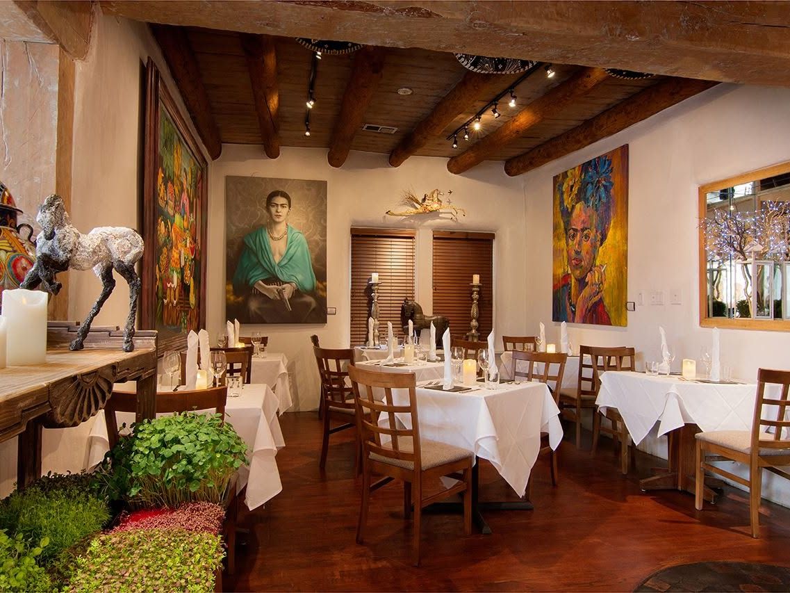 A warmly lit dining room features exposed viga ceiling beams, white adobe walls, and crisp white-linen tables set with candles and folded napkins. Vivid portraits of Frida Kahlo and sculptural artwork give the intimate space a distinctly artistic Southwestern character.