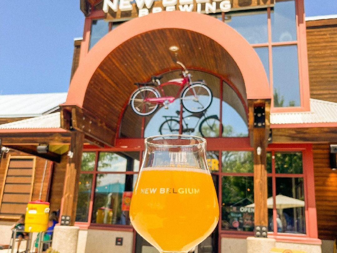 A bright glass of hazy beer is held up in front of the iconic New Belgium Brewing entrance in Asheville. This spot is perfect for enjoying sunshine, mountain views, and some of the smoothest beers in town.