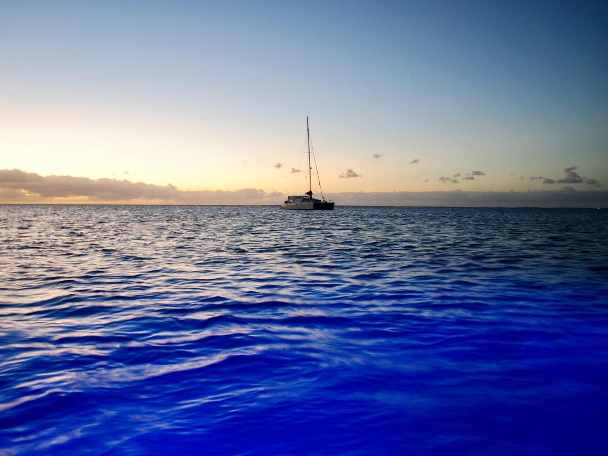 Sail boat on ocean water with blue light underneath ocean water and sunset in the background