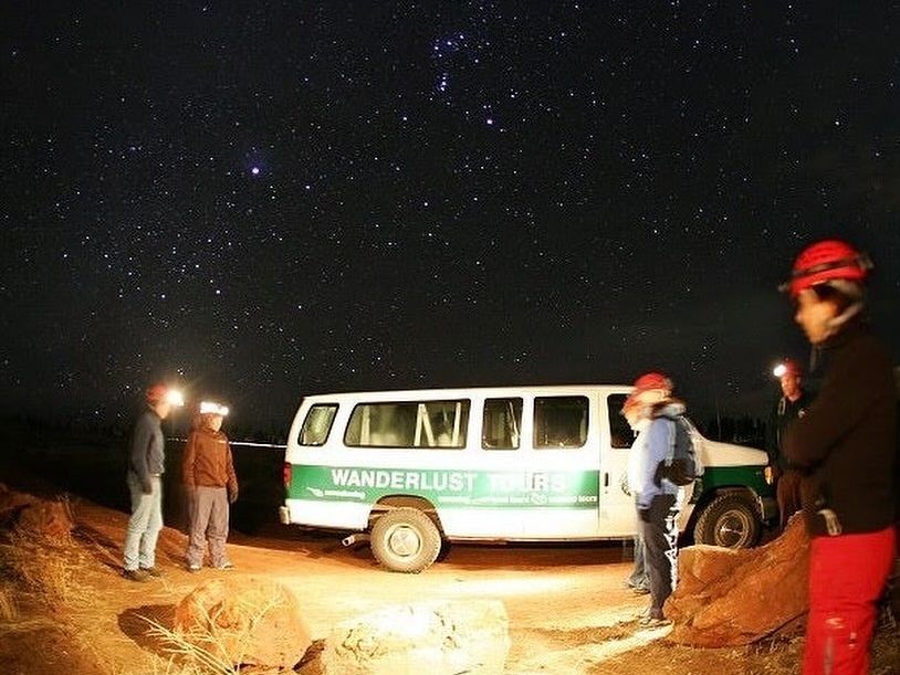 Adventurers on a nighttime cave tour under a star-filled sky, equipped with helmets and headlamps near a tour van.