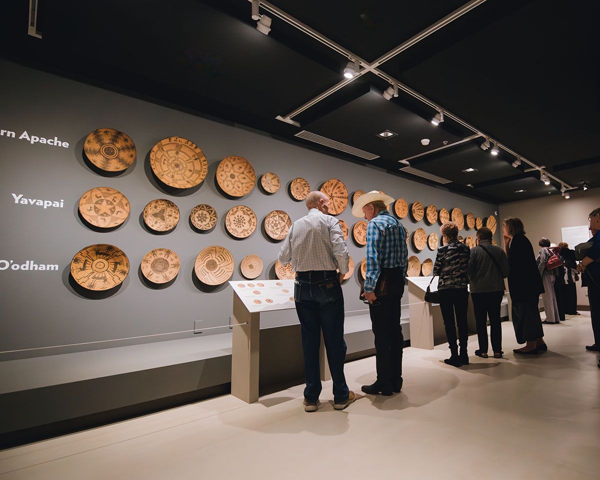 Museum visitors study rows of beautifully crafted woven baskets displayed along a soft gray wall. The detailed patterns highlight Native American artistry, while spotlights make each piece stand out. The quiet space invites people to slow down and appreciate the history behind the work.