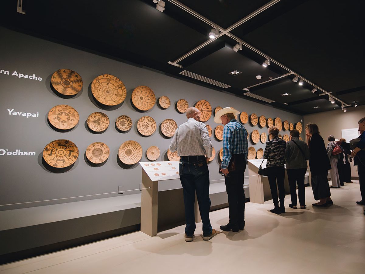Museum visitors study rows of beautifully crafted woven baskets displayed along a soft gray wall. The detailed patterns highlight Native American artistry, while spotlights make each piece stand out. The quiet space invites people to slow down and appreciate the history behind the work.