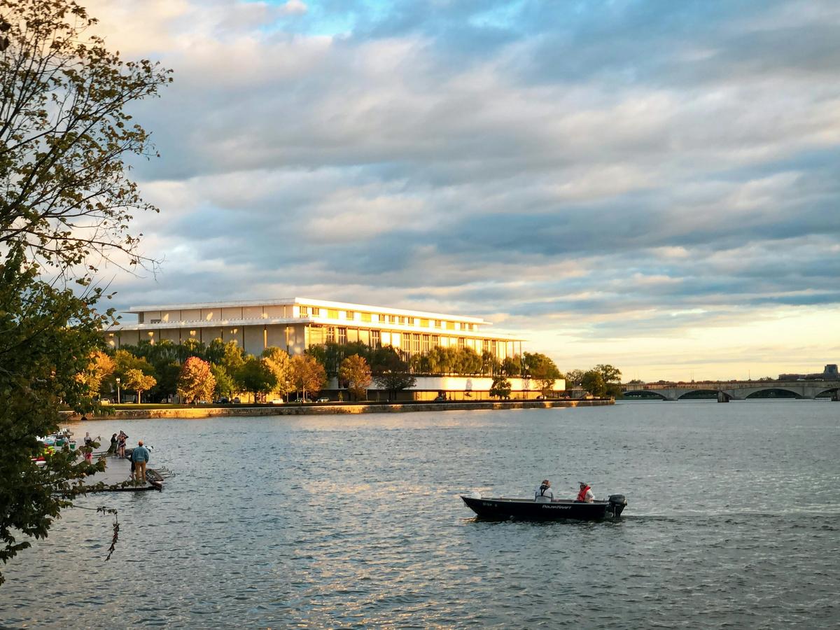 A small boat cruises along the Potomac River at sunset, with people fishing on a nearby dock and the Kennedy Center glowing in the golden hour light.