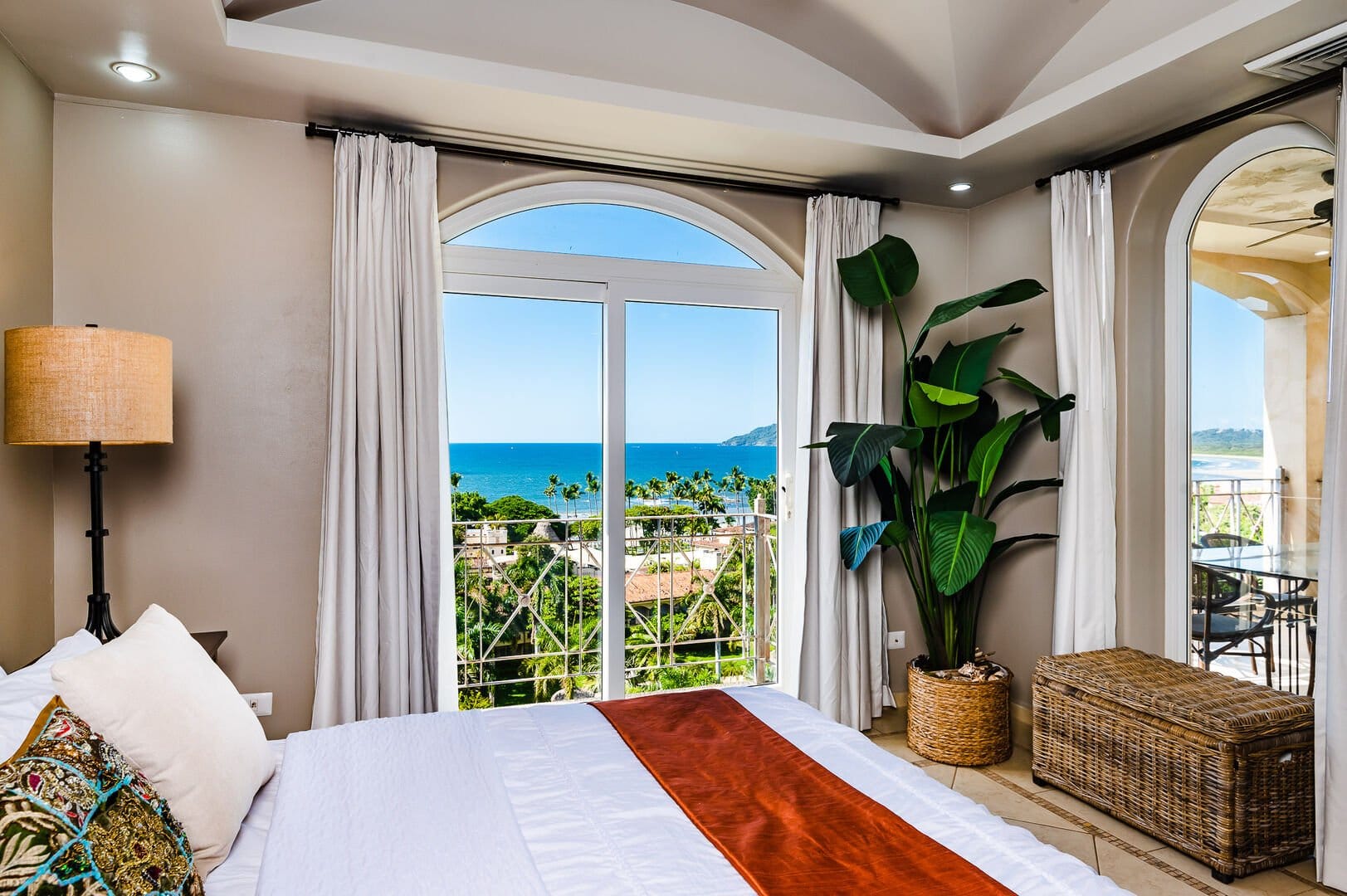 Bedroom with a large window and balcony doors overlooking the ocean, featuring neutral decor, a tropical plant, and wicker accents.