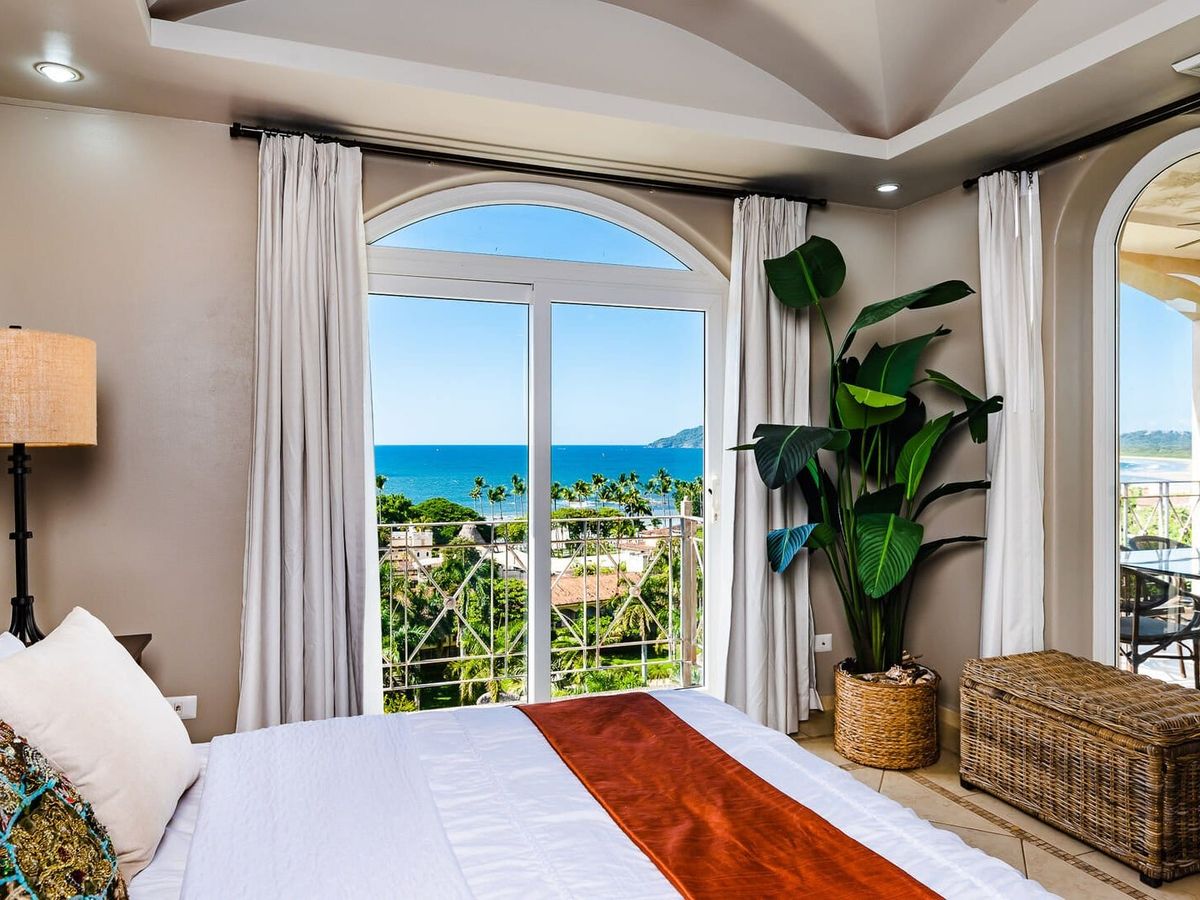 Bedroom with a large window and balcony doors overlooking the ocean, featuring neutral decor, a tropical plant, and wicker accents.