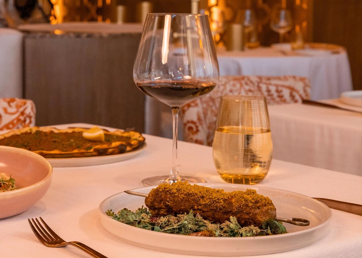A beautifully set dining table at Habibi Restaurant in Los Cabos showcases refined elegance and warm ambiance. A gourmet dish, paired with a glass of red wine, sits atop a white linen table surrounded by golden light, inviting guests to savor a luxurious culinary experience.