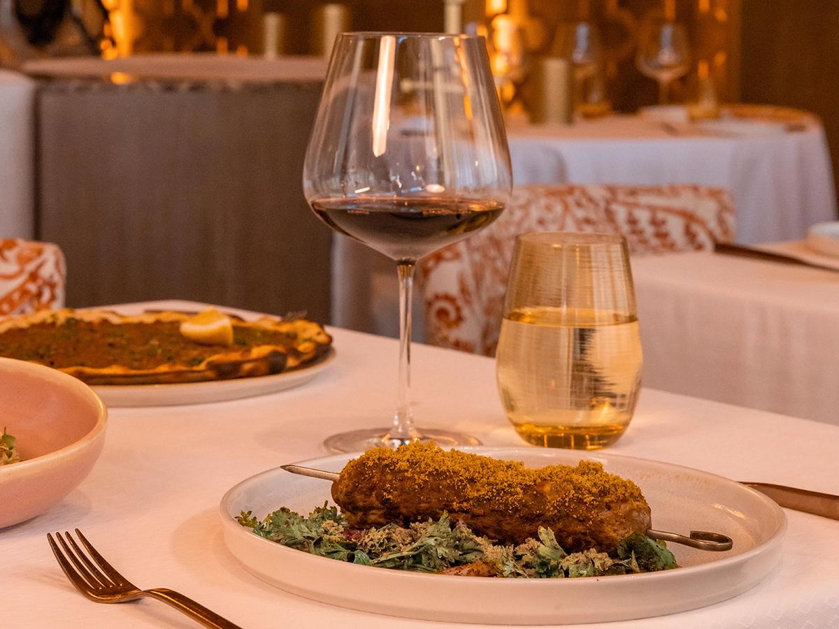 A beautifully set dining table at Habibi Restaurant in Los Cabos showcases refined elegance and warm ambiance. A gourmet dish, paired with a glass of red wine, sits atop a white linen table surrounded by golden light, inviting guests to savor a luxurious culinary experience.