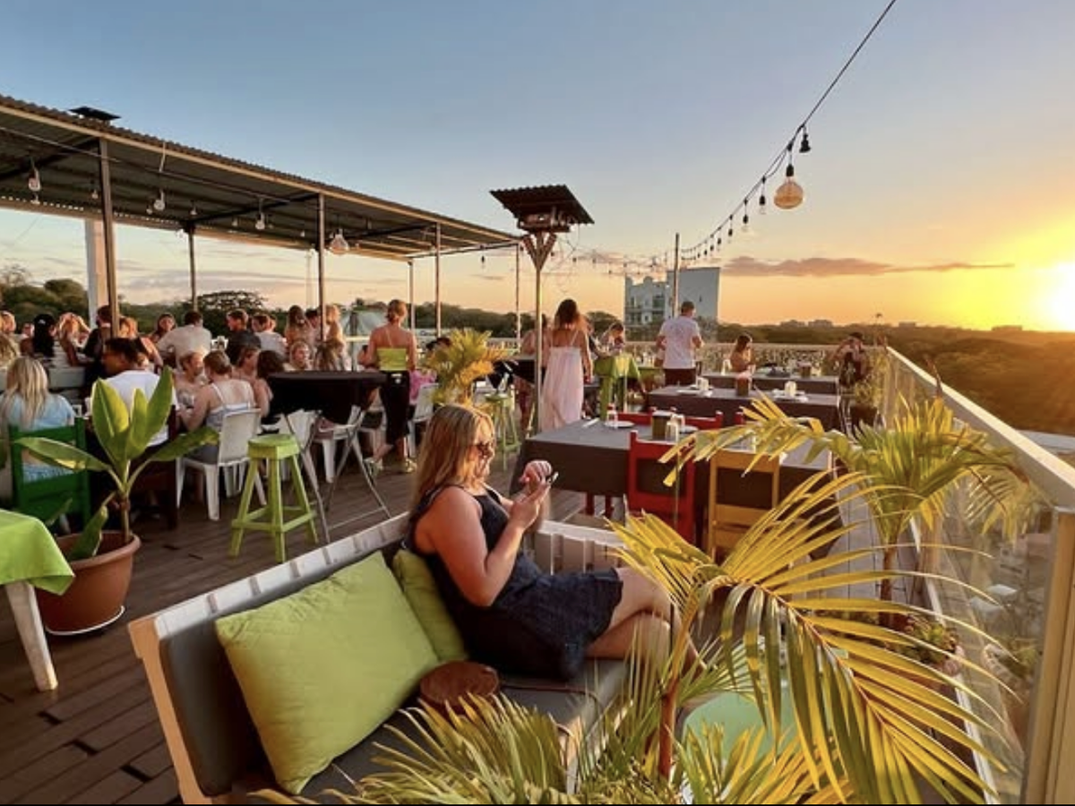 Lively rooftop restaurant at sunset, with people dining under string lights and tropical plants, overlooking a golden horizon