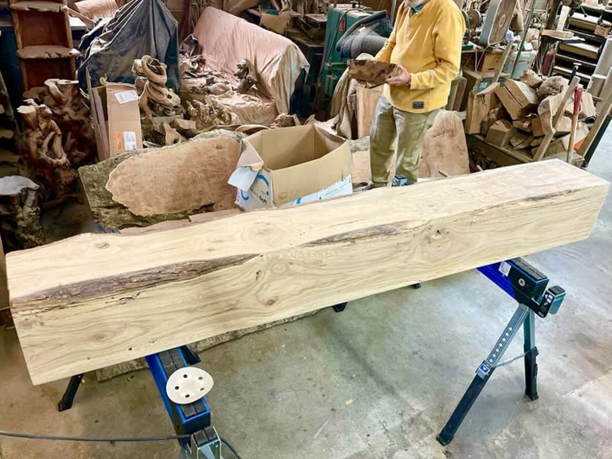 A large, freshly cut wooden beam with visible grain and knots rests on sawhorses in a cluttered woodworking shop.