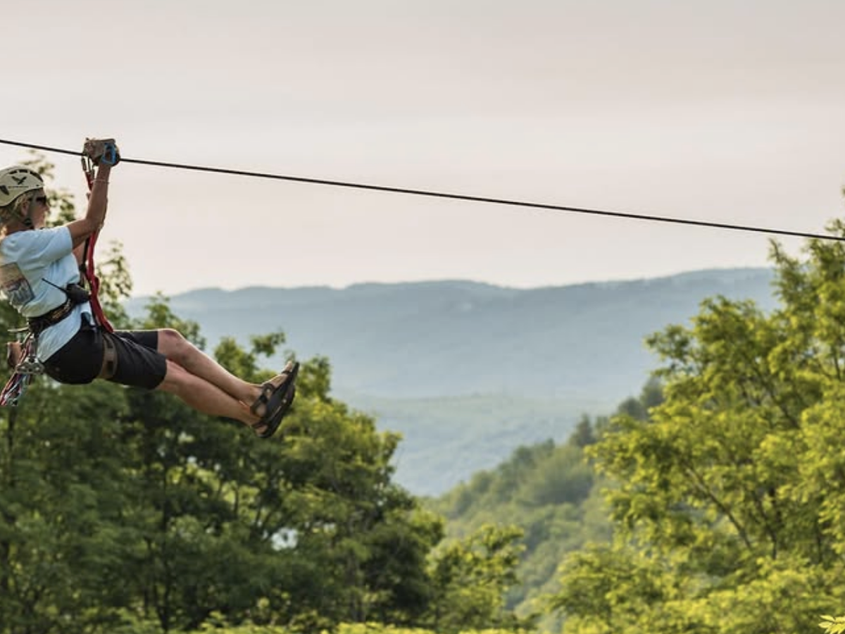 A person ziplines through the treetops with forested mountains in the background and soft afternoon light illuminating the scene.