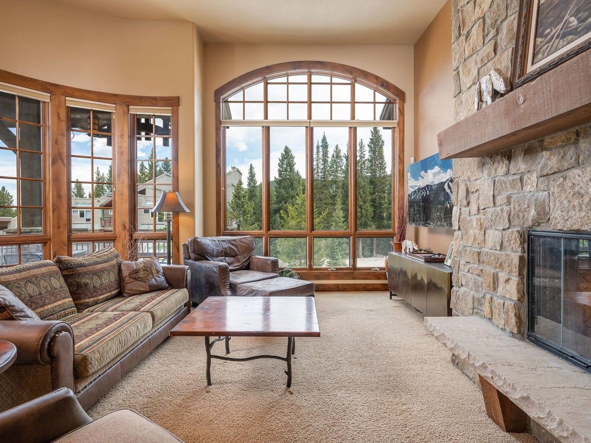 A warm and inviting mountain living room with large arched windows showcasing forest views, a stone fireplace with a wooden mantle, cozy leather and fabric seating, and rustic wood accents throughout.