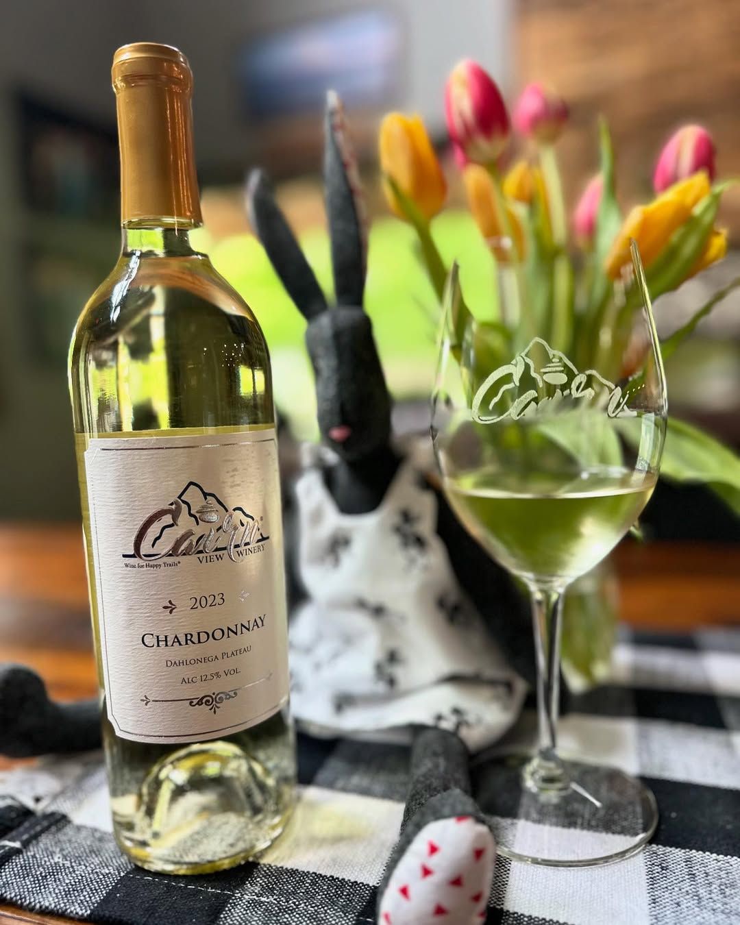 A bottle of Cairn View Chardonnay sits beside a glass of white wine on a cozy table with fresh tulips in the background. The setup feels warm and homelike, perfect for a quiet tasting moment. It captures a simple but charming wine experience.
