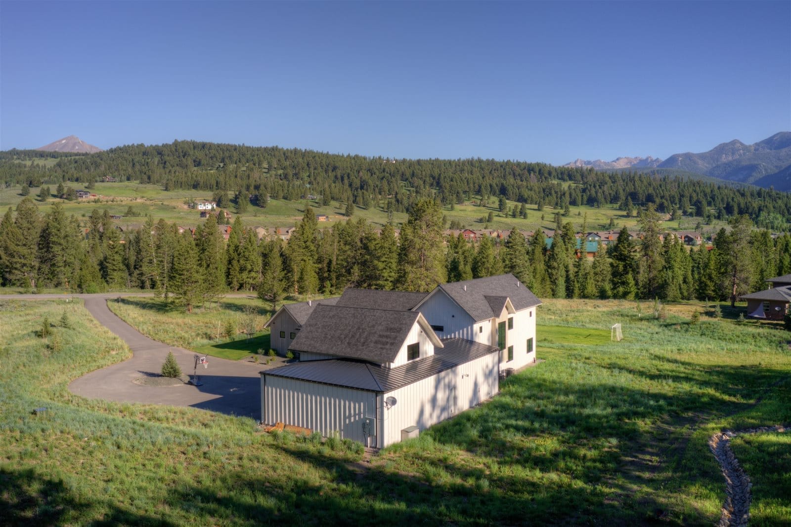 A modern farmhouse-style home with a large attached garage sits in a wide open meadow, surrounded by pine trees and mountain views under a clear blue sky.
