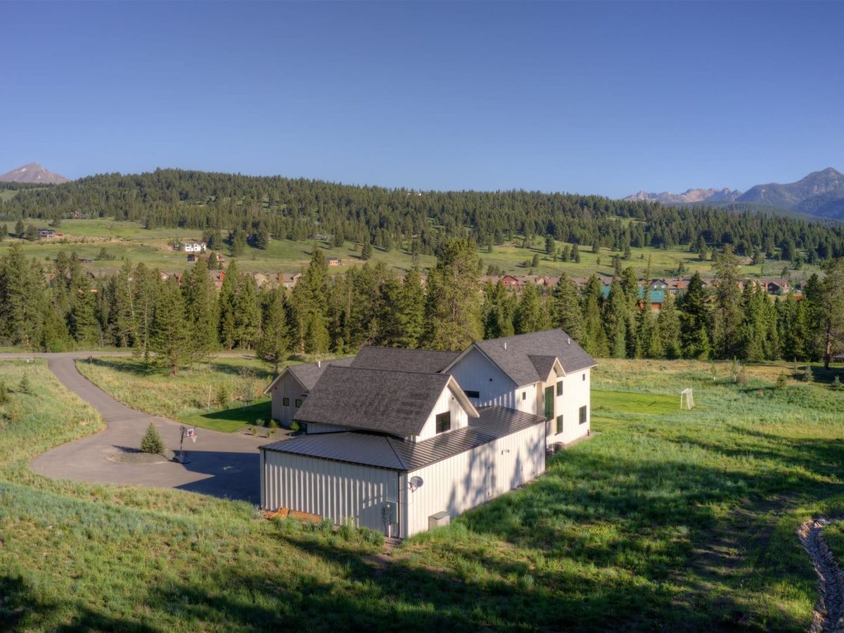 A modern farmhouse-style home with a large attached garage sits in a wide open meadow, surrounded by pine trees and mountain views under a clear blue sky.