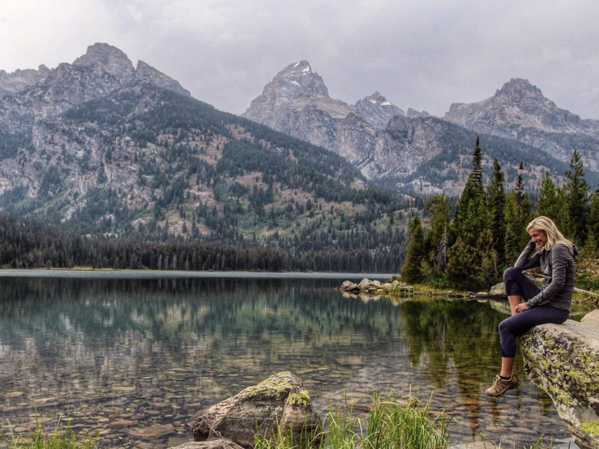 Woman sitting on a rock by a clear mountain lake, smiling with her feet dangling above the water, surrounded by evergreen trees and dramatic alpine peaks.