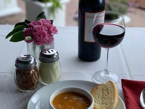 A bowl of soup served with fresh bread and a glass of red wine on a cozy table setting. Vicini’s Italian Restaurant & Pizzeria offers homestyle Italian cooking in a warm, intimate atmosphere.