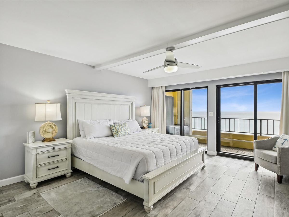 A serene coastal-style bedroom with a white wooden bed, matching nightstands, a modern ceiling fan, and sliding glass doors that open to a private balcony with ocean views.