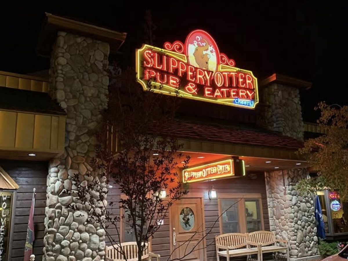 The exterior of Slippery Otter Pub & Eatery at night, featuring a glowing neon sign above a rustic stone-and-wood building with benches by the entrance.