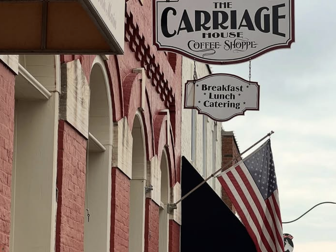 A vintage-style hanging sign for The Carriage House Coffee Shoppe advertises breakfast, lunch, and catering, mounted on a red-brick building with arched white trim and an American flag displayed nearby.