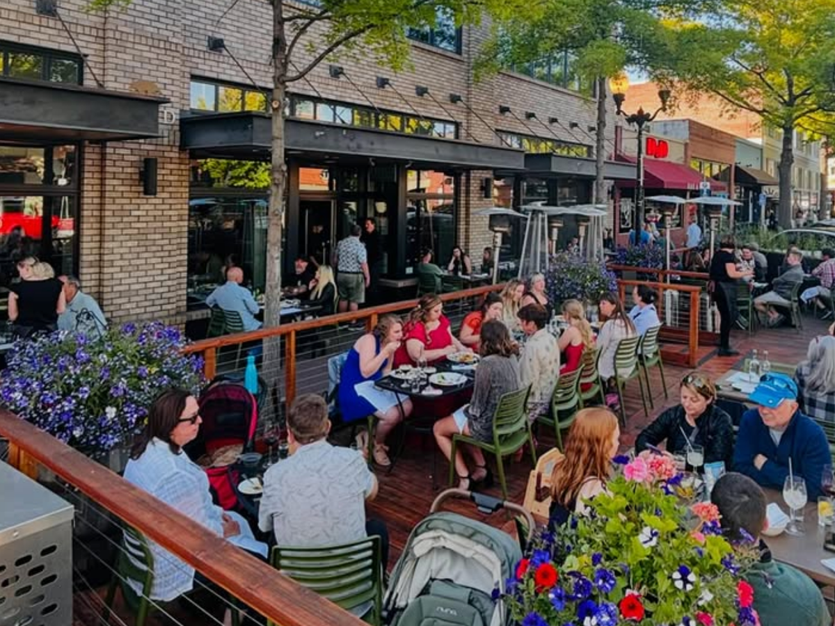Bustling outdoor dining scene with groups of people enjoying food and drinks on a lively patio lined with flowers and trees.