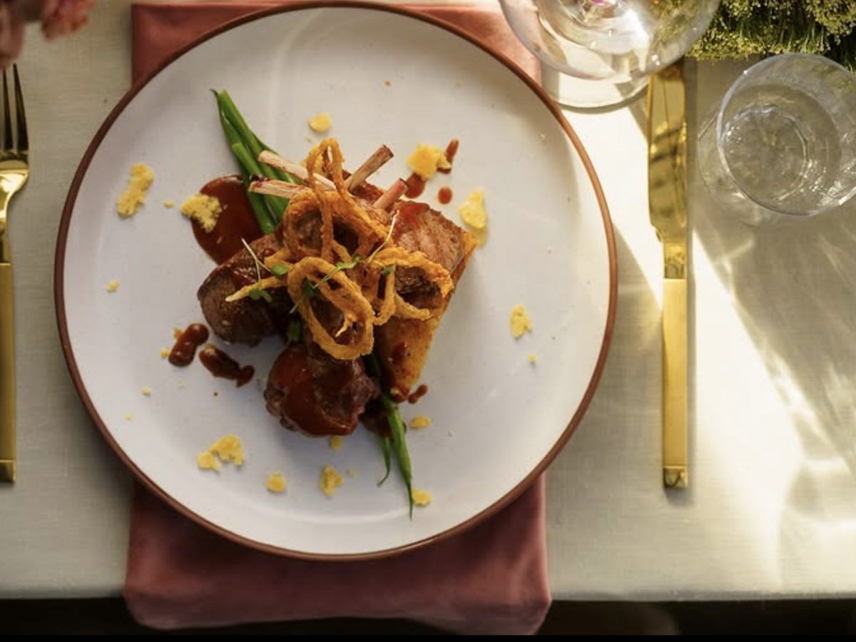 Elegant entrée featuring rack of lamb atop green beans and polenta, finished with a savory demi-glace and crispy fried onions, served on a white plate with golden cutlery.