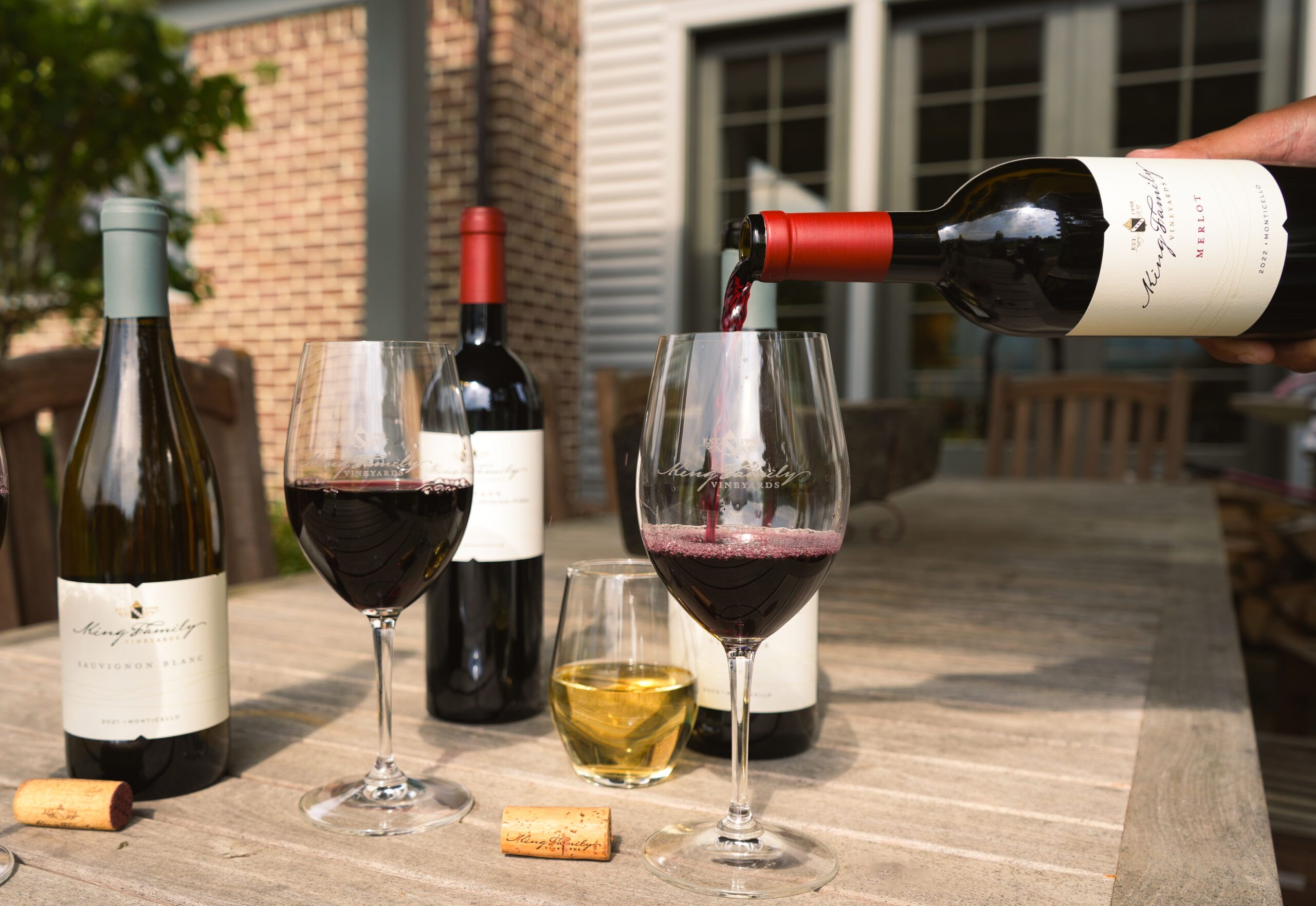 A pour of red wine fills a glass beside bottles of King Family Vineyards wine on a sunny patio table. The elegant presentation and soft lighting showcase the winery’s rich tradition and scenic Monticello countryside.
