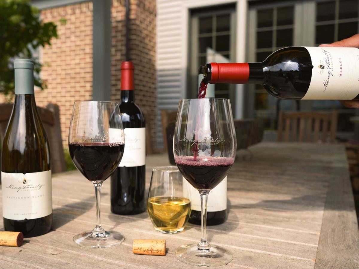 A pour of red wine fills a glass beside bottles of King Family Vineyards wine on a sunny patio table. The elegant presentation and soft lighting showcase the winery’s rich tradition and scenic Monticello countryside.
