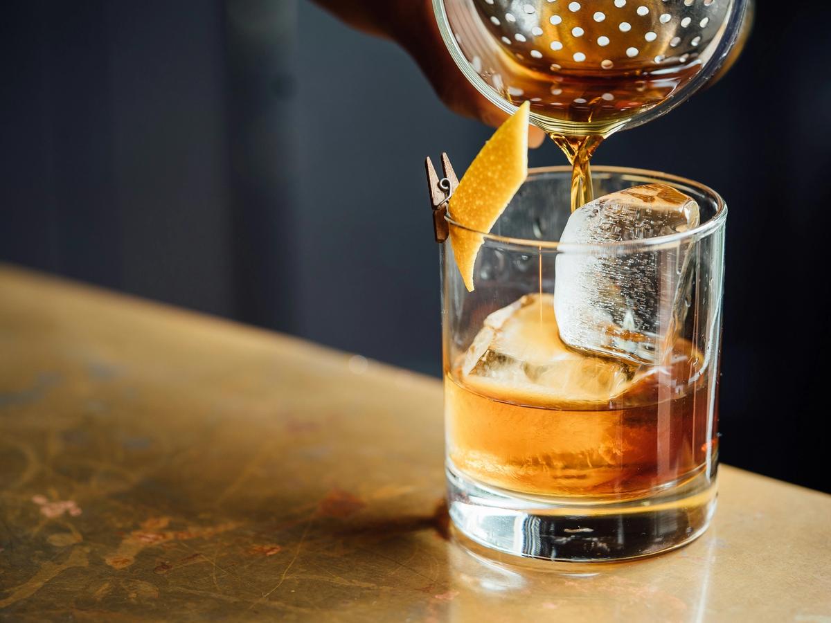 A close-up of a bartender pouring a golden cocktail over a large ice cube into a rocks glass, garnished with a twist of citrus peel clipped to the rim.