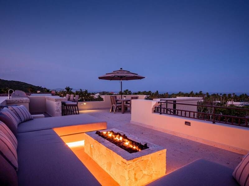 Rooftop terrace at twilight featuring a modern stone fire pit, cozy sectional seating, dining table with umbrella, and views of tropical greenery and distant hills.