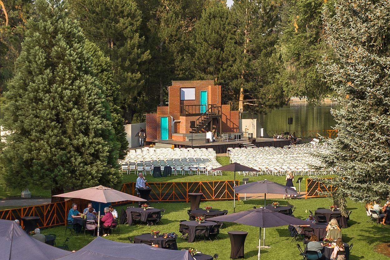 Rows of white chairs face an outdoor stage set with a brick backdrop and turquoise doors surrounded by tall pine trees. Guests relax under umbrellas before showtime, creating a peaceful and inviting atmosphere at Bend’s Theatre in the Park.