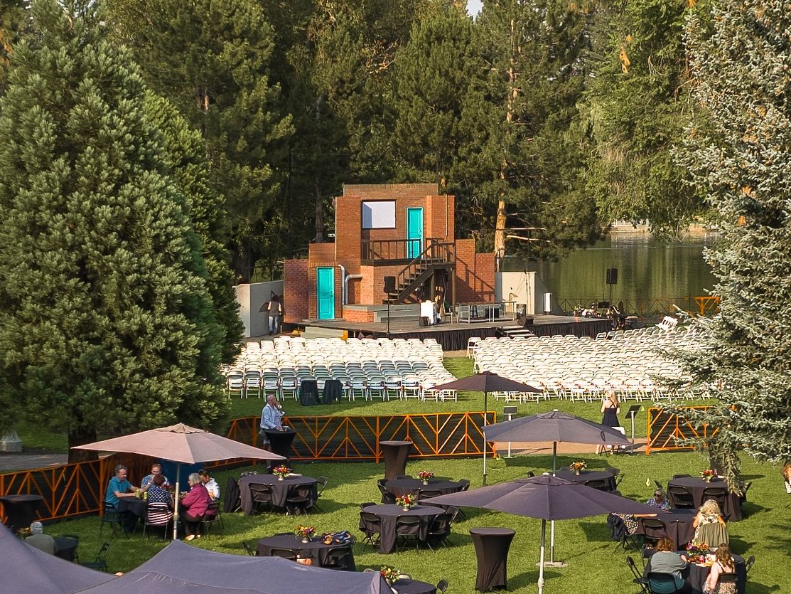 Rows of white chairs face an outdoor stage set with a brick backdrop and turquoise doors surrounded by tall pine trees. Guests relax under umbrellas before showtime, creating a peaceful and inviting atmosphere at Bend’s Theatre in the Park.