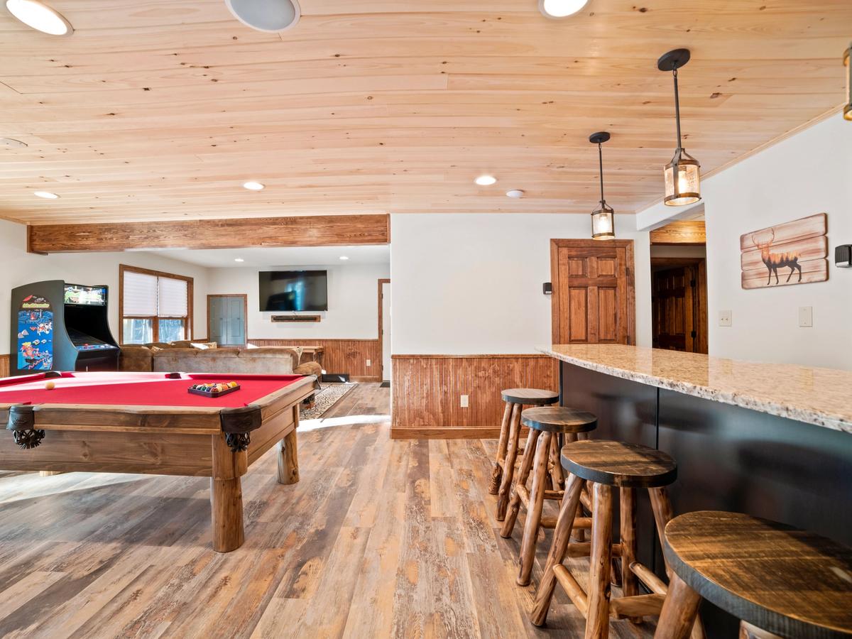 Rustic game room with a red felt pool table, wooden bar with stools, arcade game machine, cozy seating area, and mounted TV.