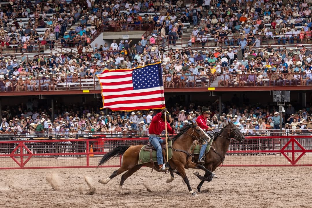 Two horseback riders gallop across the arena waving a large American flag as the crowd cheers from packed stands. The lively scene captures the patriotic energy and excitement of the Cody Frontier Festival.