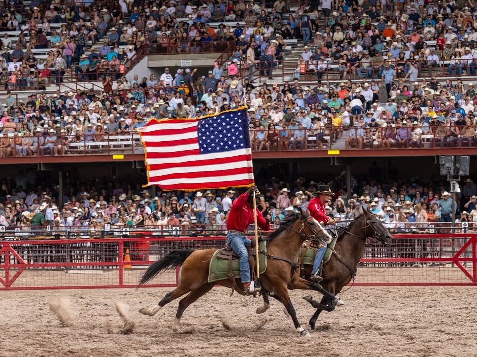Two horseback riders gallop across the arena waving a large American flag as the crowd cheers from packed stands. The lively scene captures the patriotic energy and excitement of the Cody Frontier Festival.