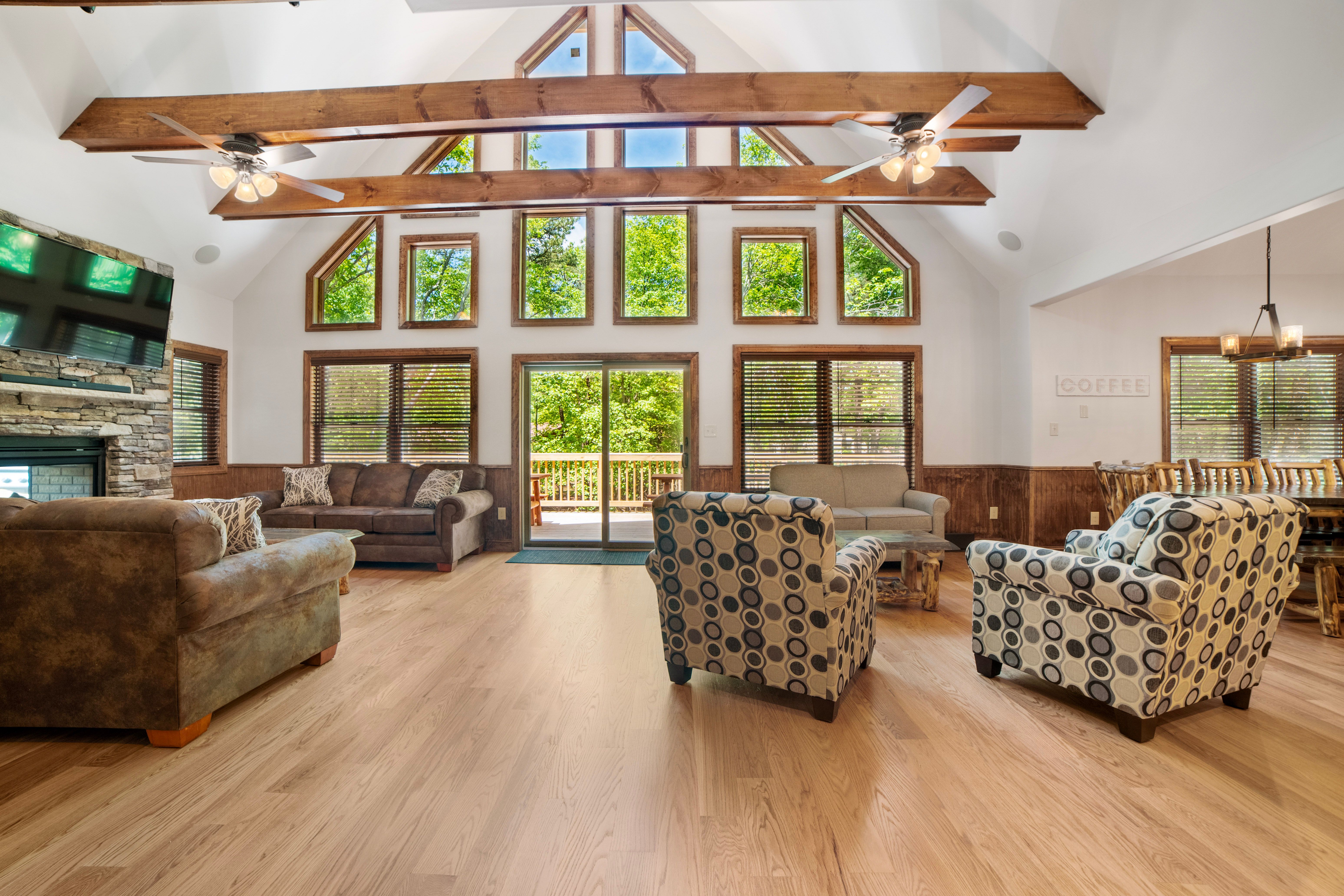 Spacious living room with vaulted ceilings, exposed wooden beams, large windows, cozy seating, a stone fireplace with a mounted TV, and views of the outdoor deck.