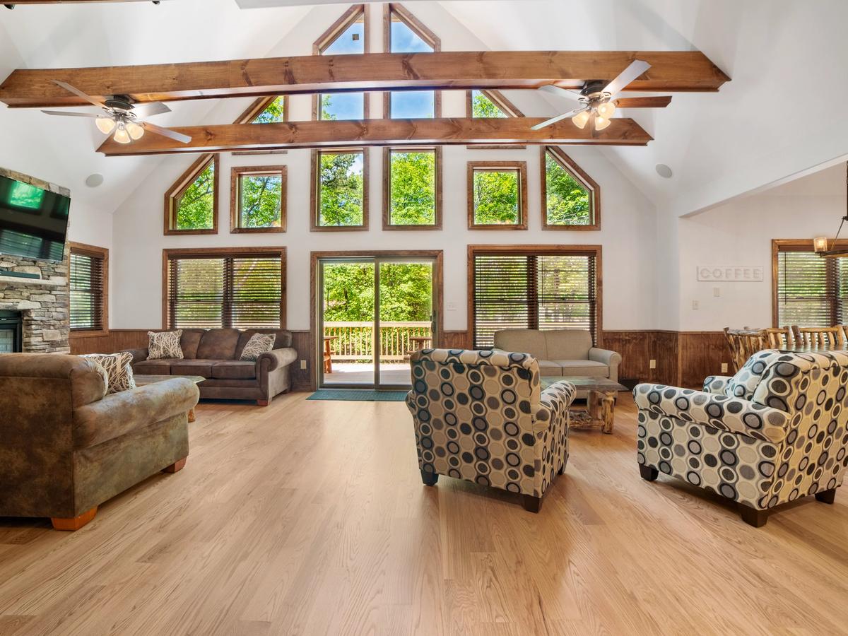 Spacious living room with vaulted ceilings, exposed wooden beams, large windows, cozy seating, a stone fireplace with a mounted TV, and views of the outdoor deck.
