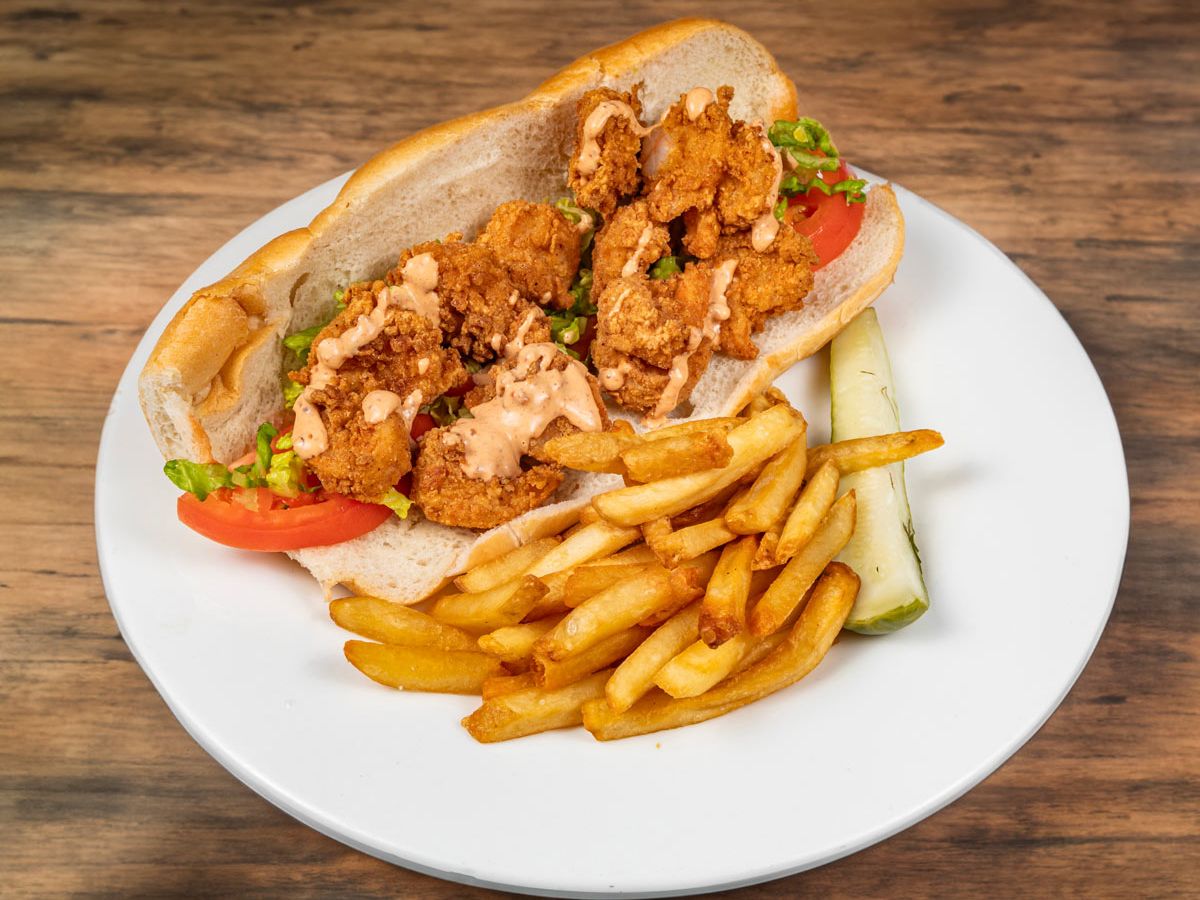 A crispy shrimp po’ boy sandwich loaded with lettuce, tomato, and sauce, served with fries and a pickle spear. A true taste of Southern comfort.