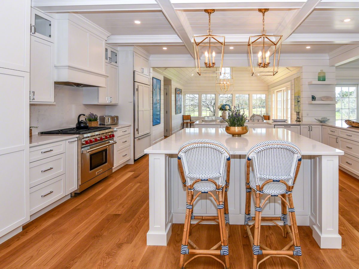 A bright, coastal-style kitchen features white cabinetry, a large center island with two woven barstools, brass pendant lighting, and wide-plank hardwood floors.