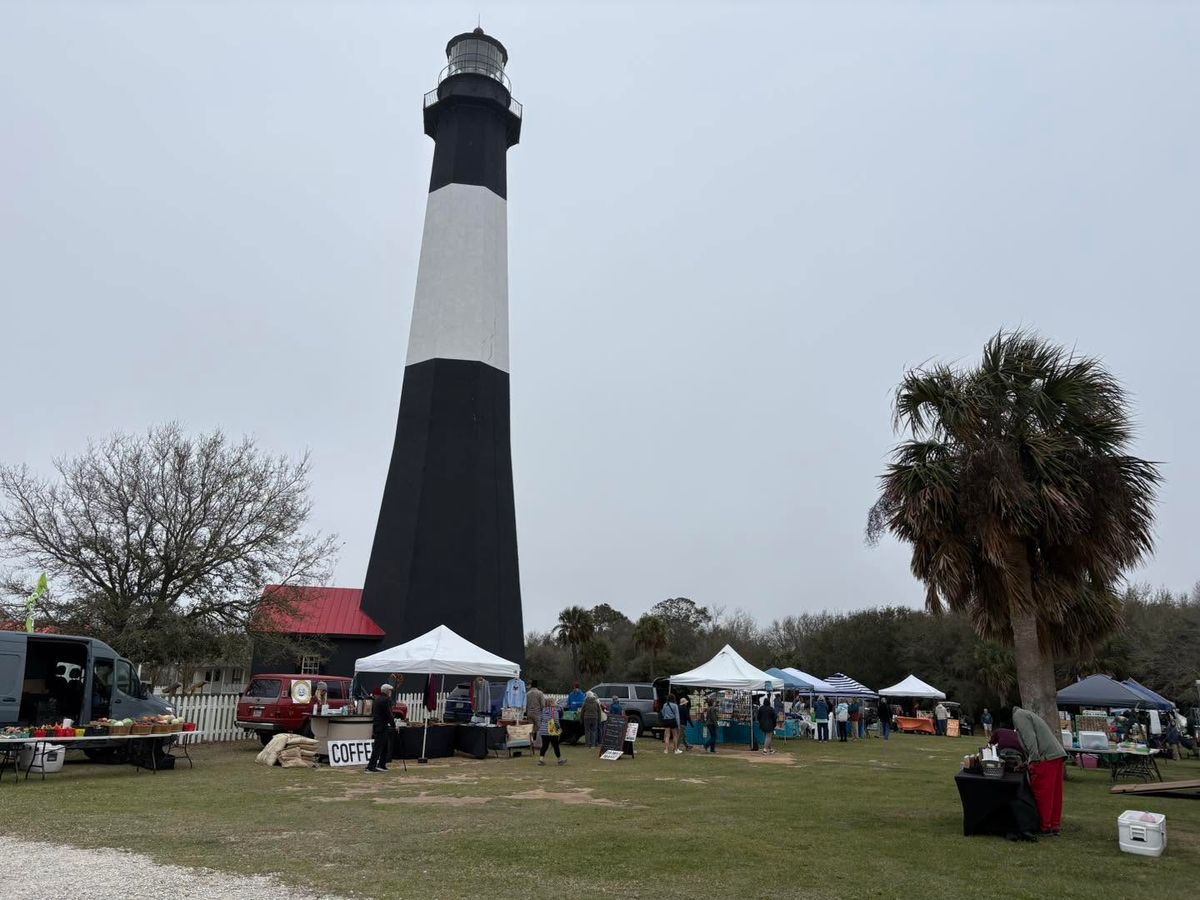  The black-and-white Tybee Island Lighthouse rises above a lively outdoor market with white vendor tents, shoppers, and a coffee stand set on a grassy field. A palm tree and bare winter trees frame the scene beneath an overcast gray sky