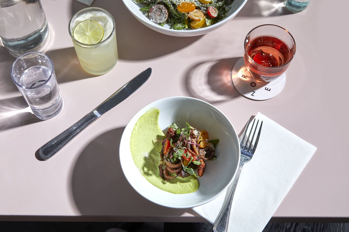 A minimalist yet refined dining setup featuring a bowl of seasonal vegetables in a light green sauce, paired with refreshing cocktails. The soft pastel tones and natural light create a calm, stylish café atmosphere.