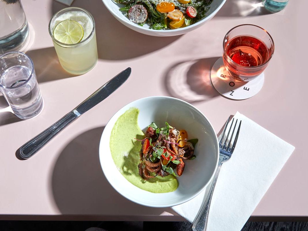 A minimalist yet refined dining setup featuring a bowl of seasonal vegetables in a light green sauce, paired with refreshing cocktails. The soft pastel tones and natural light create a calm, stylish café atmosphere.