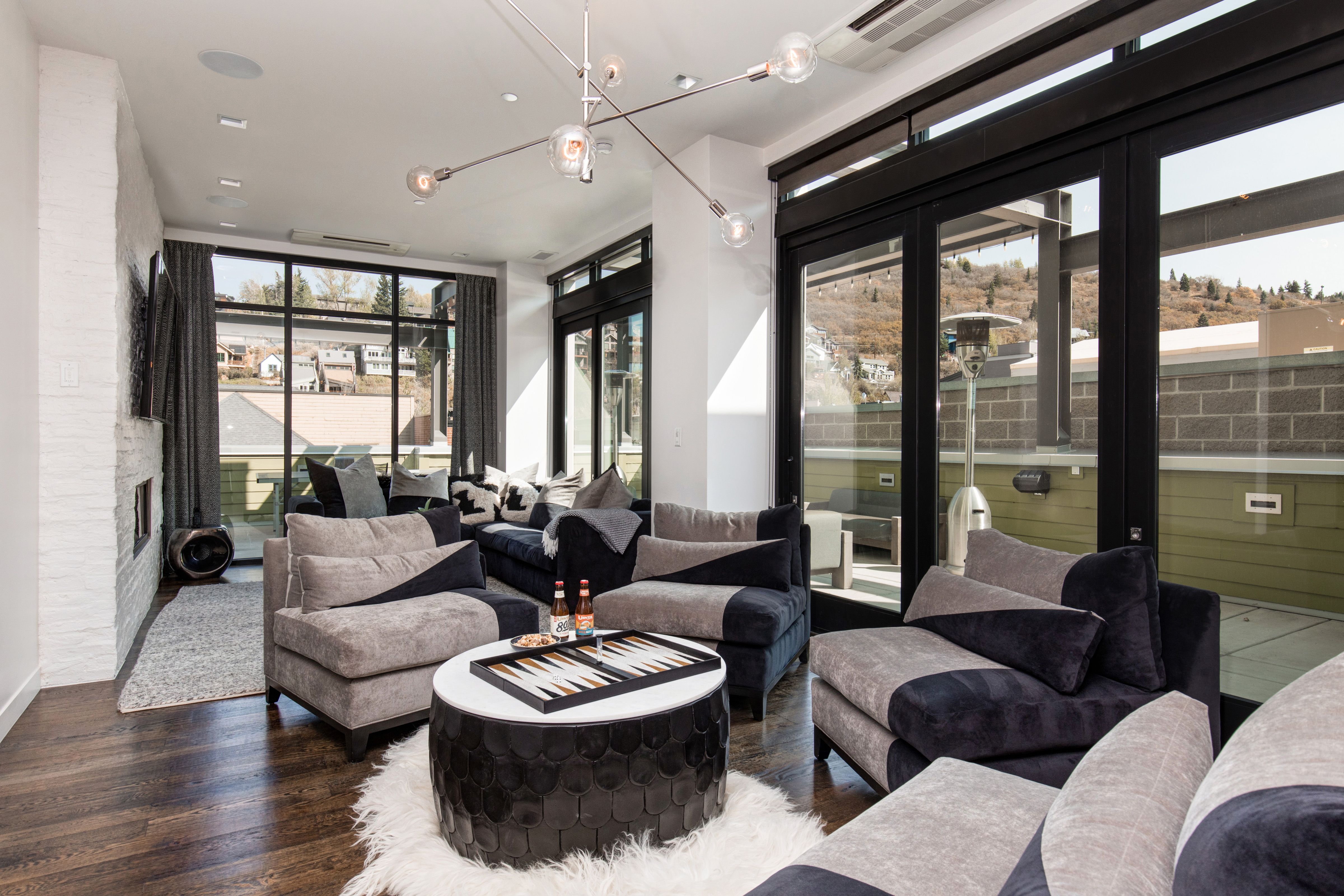 A chic, contemporary penthouse lounge with plush two-tone chairs, a round black-and-white backgammon table, modern lighting, and floor-to-ceiling windows that open to a private terrace with mountain views.