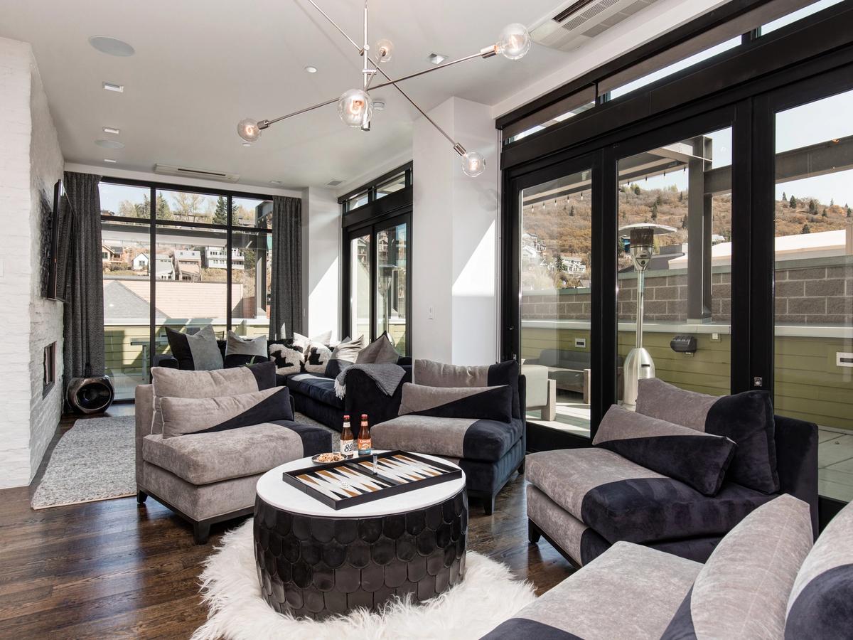 A chic, contemporary penthouse lounge with plush two-tone chairs, a round black-and-white backgammon table, modern lighting, and floor-to-ceiling windows that open to a private terrace with mountain views.