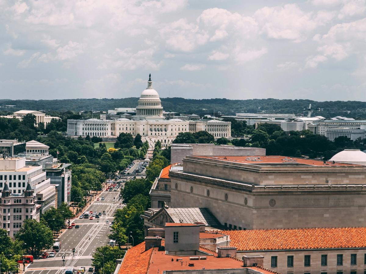 Daytime aerial view of Washington, D.C., featuring the U.S. Capitol building, tree-lined avenues, and surrounding federal architecture.