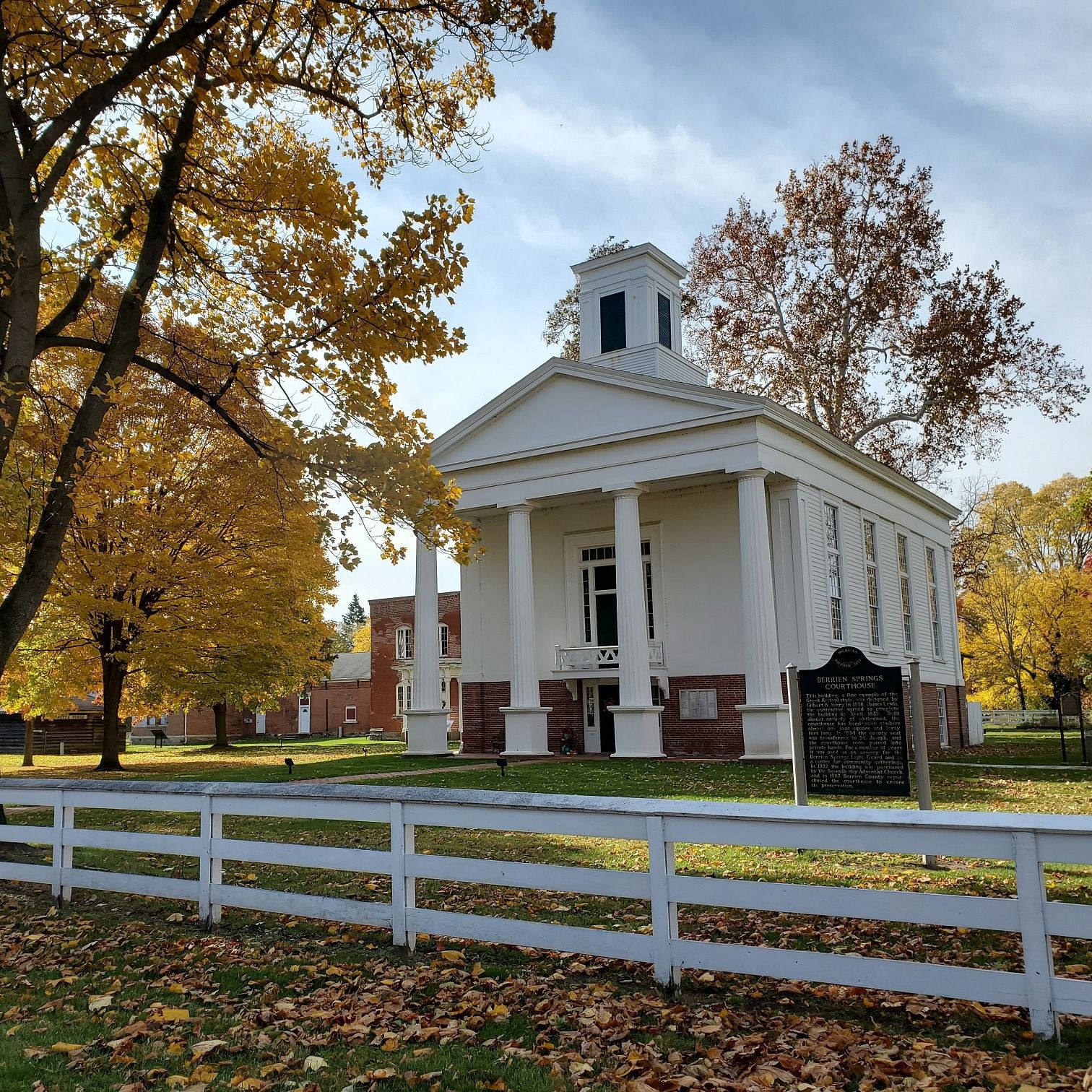 This historic courthouse stands tall among golden autumn trees, its white columns and red brick foundation glowing in the afternoon light. Fallen leaves scatter across the lawn, adding to the timeless small-town charm.