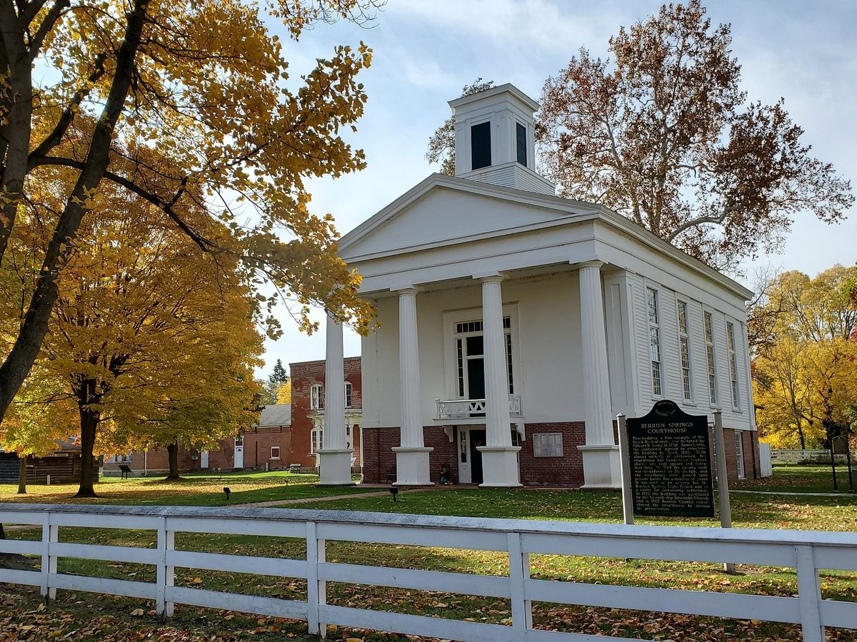 This historic courthouse stands tall among golden autumn trees, its white columns and red brick foundation glowing in the afternoon light. Fallen leaves scatter across the lawn, adding to the timeless small-town charm.