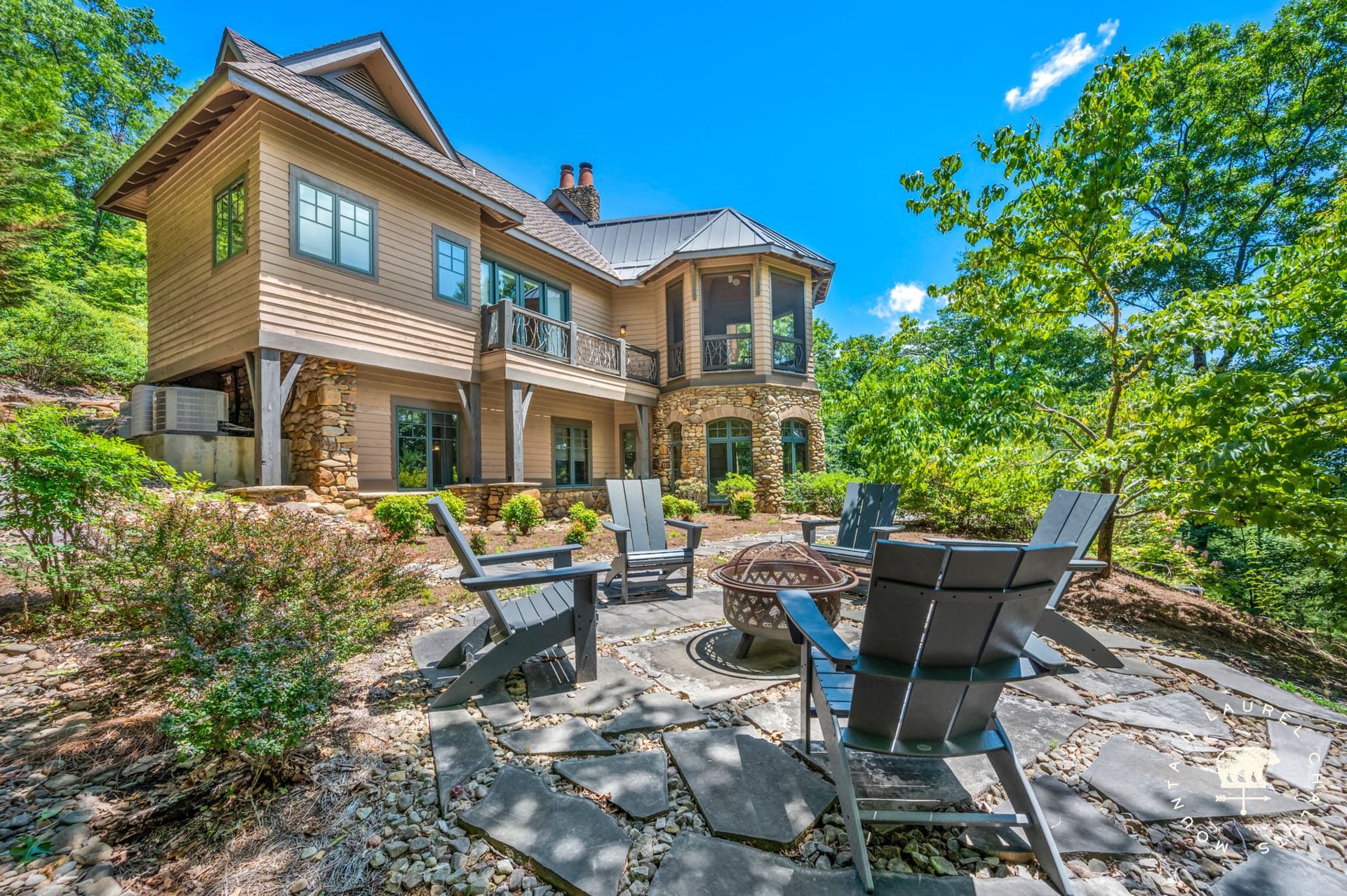 A mountain-style home with stone and wood exterior overlooking a forested setting, featuring a circular stone patio with Adirondack chairs gathered around a fire pit beneath a bright blue sky.