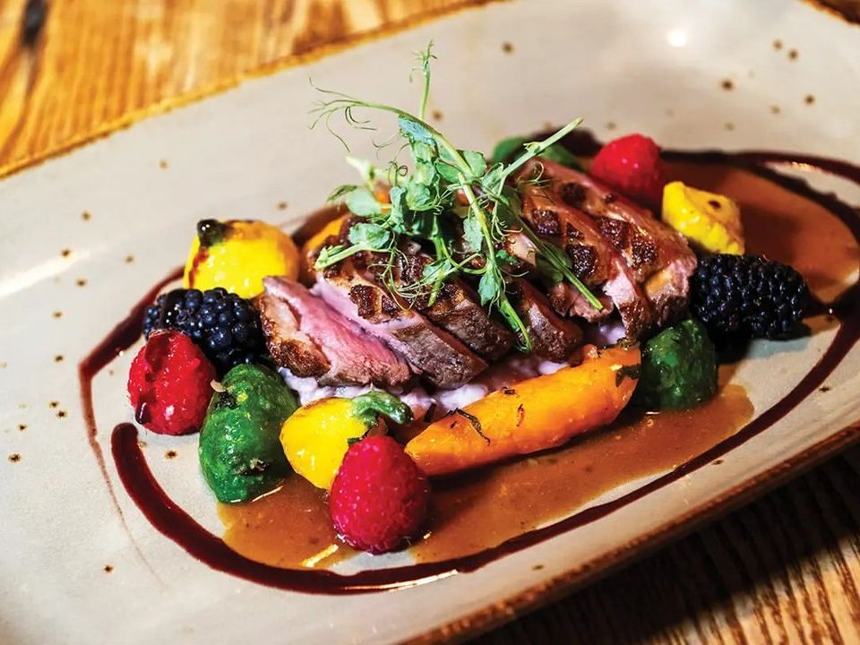 A gourmet plate of seared duck breast is served with roasted vegetables and fresh berries, finished with a rich sauce. The dish reflects the refined, farm-to-table flavors Magnolias at the Mill is known for.