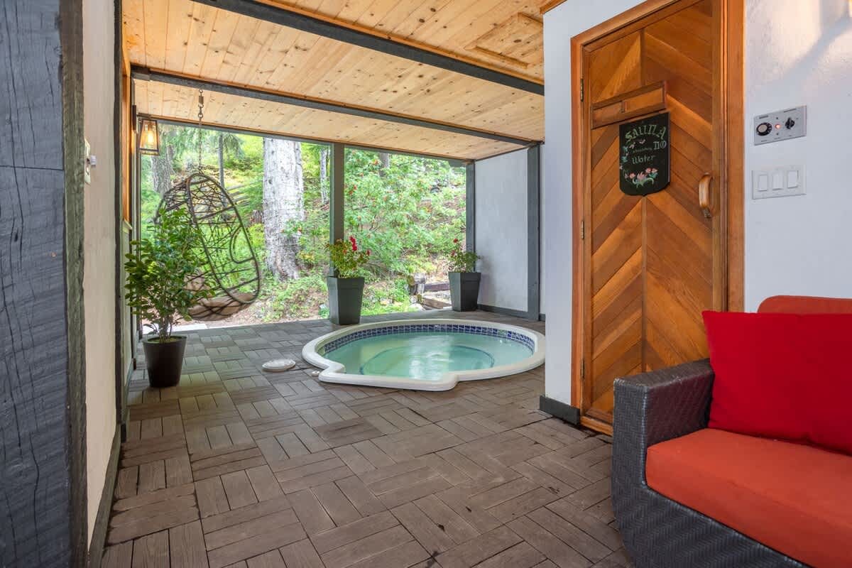 Covered outdoor spa area with a built-in circular hot tub, wood-paneled sauna door, hanging egg chair, and large screened opening to a lush wooded setting.