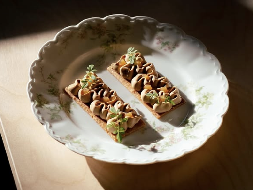 Two rectangular crackers topped with piped savory mousse and garnished with microgreens, served on a delicate floral china plate in warm, natural lighting.